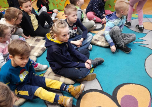 Przedszkolaki siedzą na dywanie i słuchają pani bibliotekarki